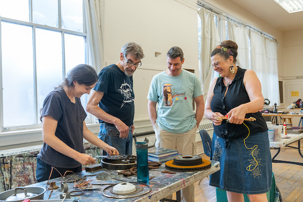 A group of 4 artists stands around a table, smiling and looking down at partially-finished assemblage sculpture on the table.